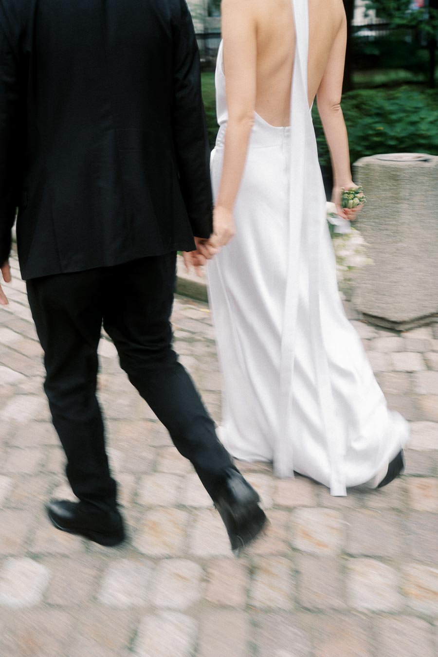 A couple walking hand in hand on a cobblestone path, with the bride in an elegant open-back white gown and the groom in a black suit, symbolizing a romantic wedding moment.