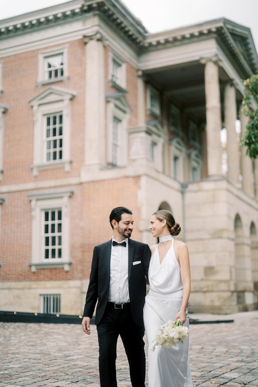 A couple in elegant wedding attire smiling at each other while strolling in front of a historic building with classical architecture.