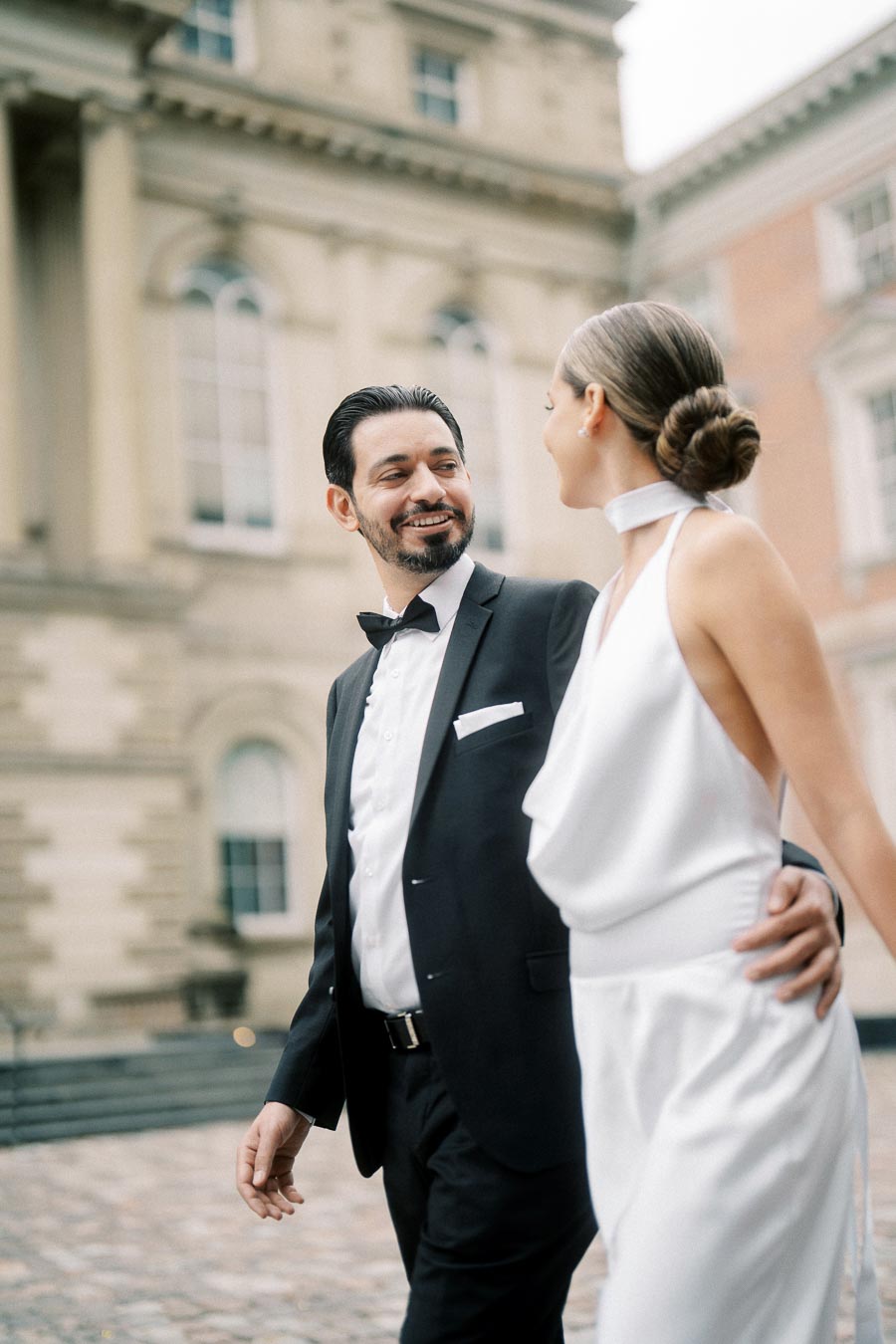 A couple elegantly dressed in formal attire, walking outside a historic building with large arched windows. The man wears a black tuxedo, and the woman dons a white gown, both smiling at each other.