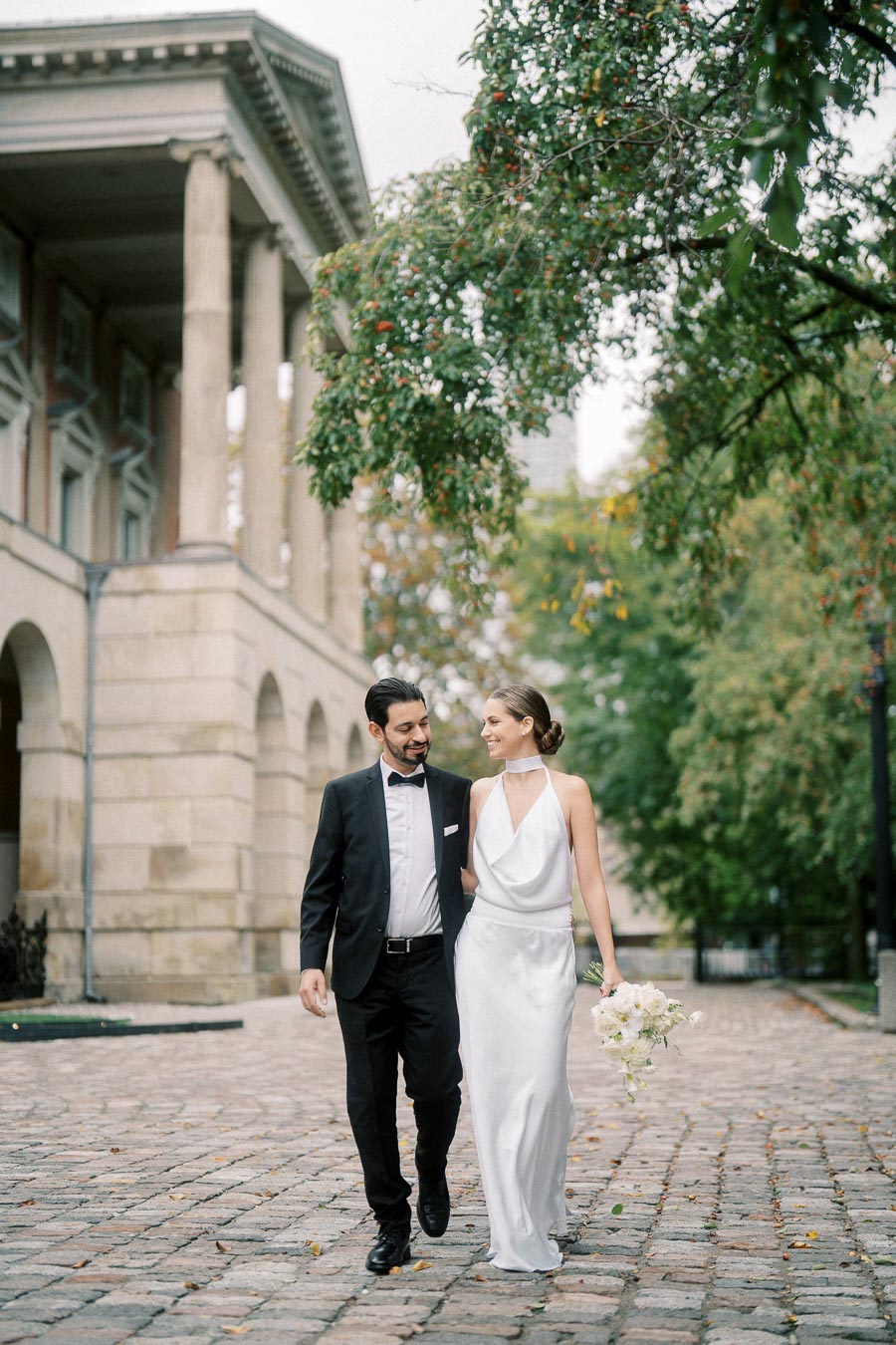 Bride and groom walking on cobblestone path under lush green trees outside a historic building, with the bride in a white gown holding a bouquet.