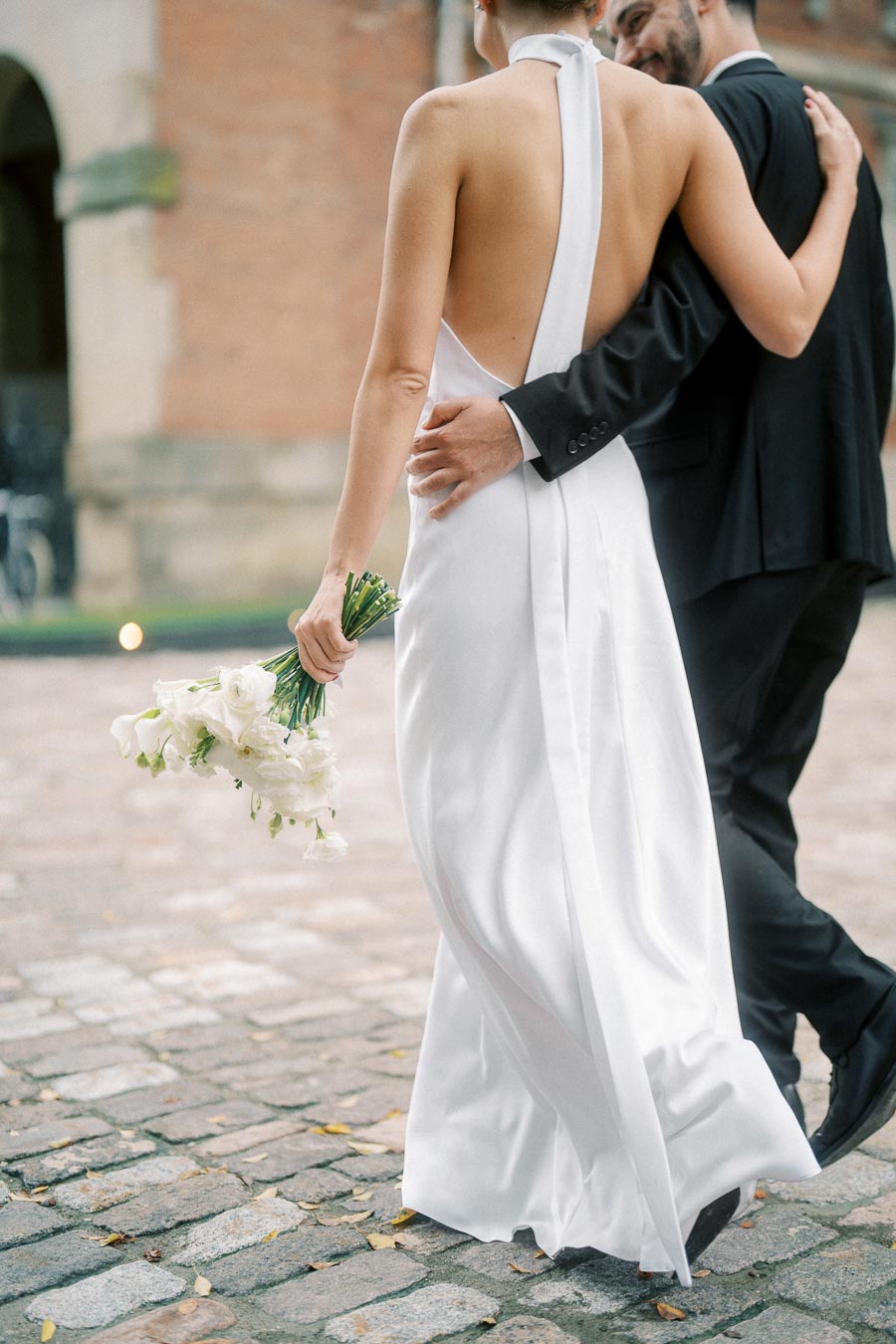 Back view of a couple walking on cobblestone in elegant wedding attire, with the bride holding a bouquet of white flowers.