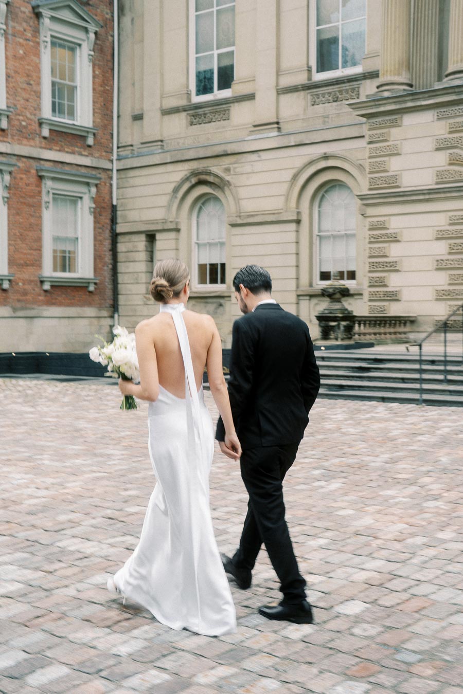 Elegant couple walking in courtyard, bride wearing a sleek white dress holding a bouquet, groom in a black suit; historic building facade in the background.