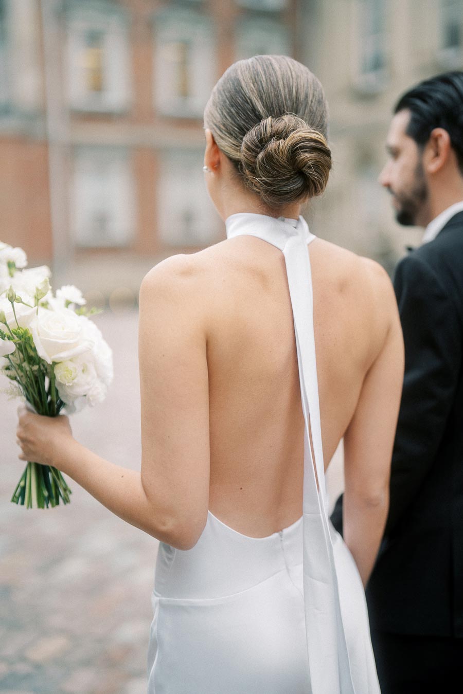 Elegant bride in a backless halterneck gown holding a bouquet of white roses, standing with a groom in a formal suit against an urban backdrop.