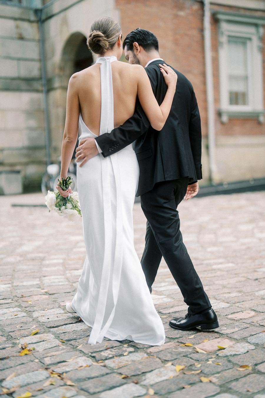 Rear view of a newlywed couple walking arm in arm on cobblestone street, the bride in an elegant backless white dress holding a bouquet, and the groom in a classic black suit, symbolizing romance and celebration in a historic urban setting.