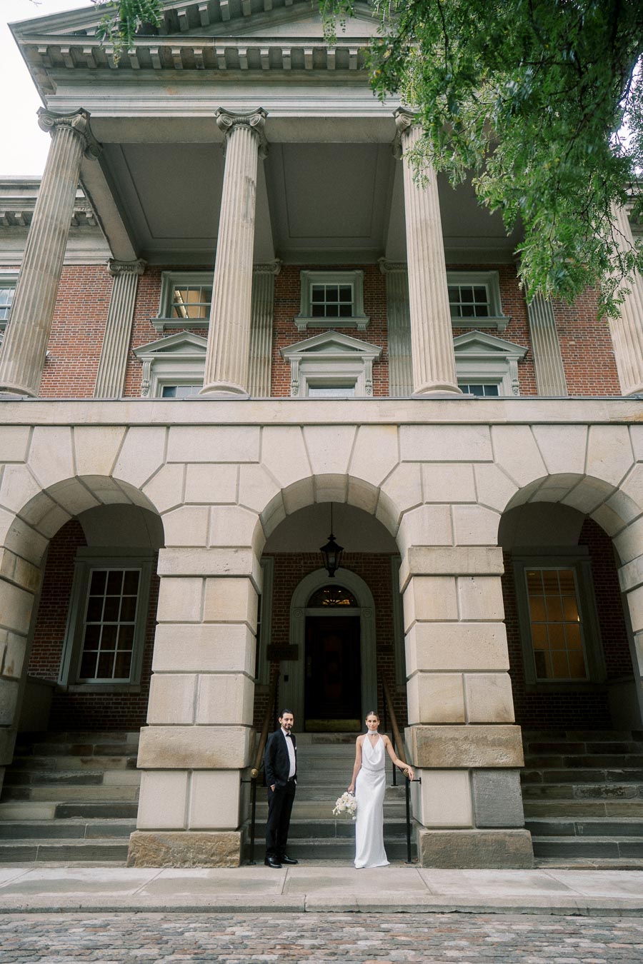 Elegant couple posing in front of a historic building with grand columns and arches, featuring a brick facade and classic architectural details, surrounded by greenery.