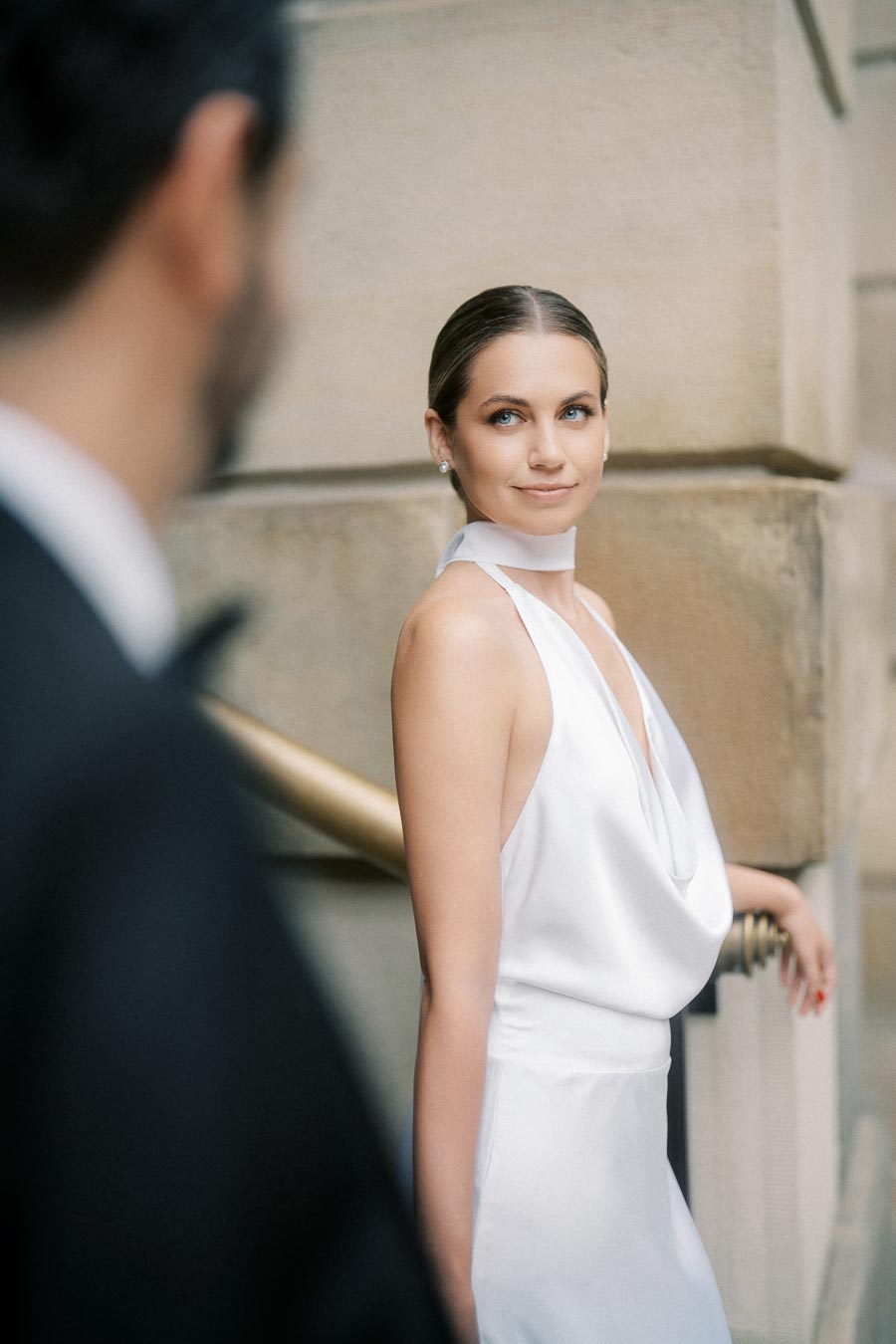 Elegant woman in a white dress smiling, leaning against a beige stone wall, with an out-of-focus person in a tuxedo in the foreground. Romantic fashion scene.
