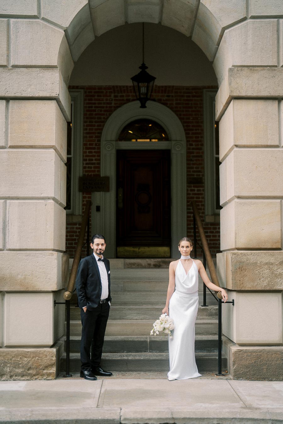 A bride in an elegant white satin gown holds a bouquet while standing next to a groom in a black tuxedo. They pose in front of a historic brick building with an arched entrance, creating a timeless wedding photo.