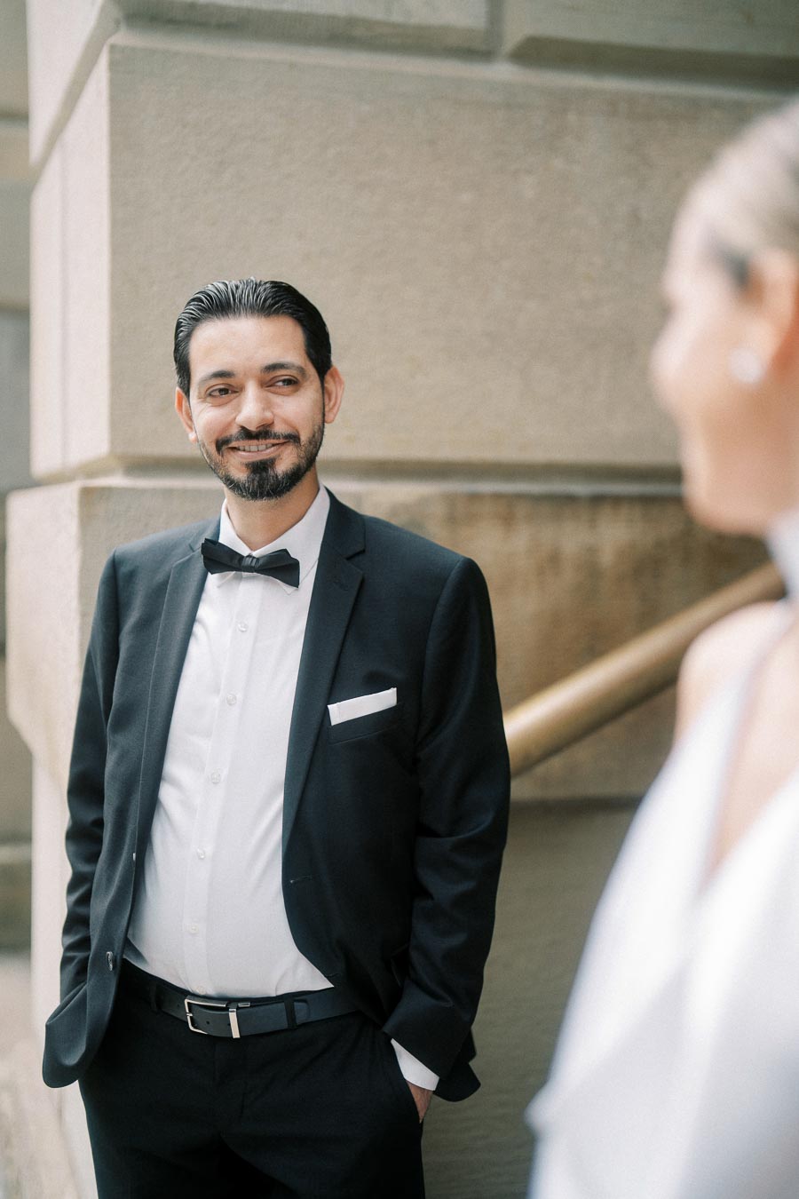 Man in a formal black suit with a bow tie smiling while standing outdoors, engaging with another person.