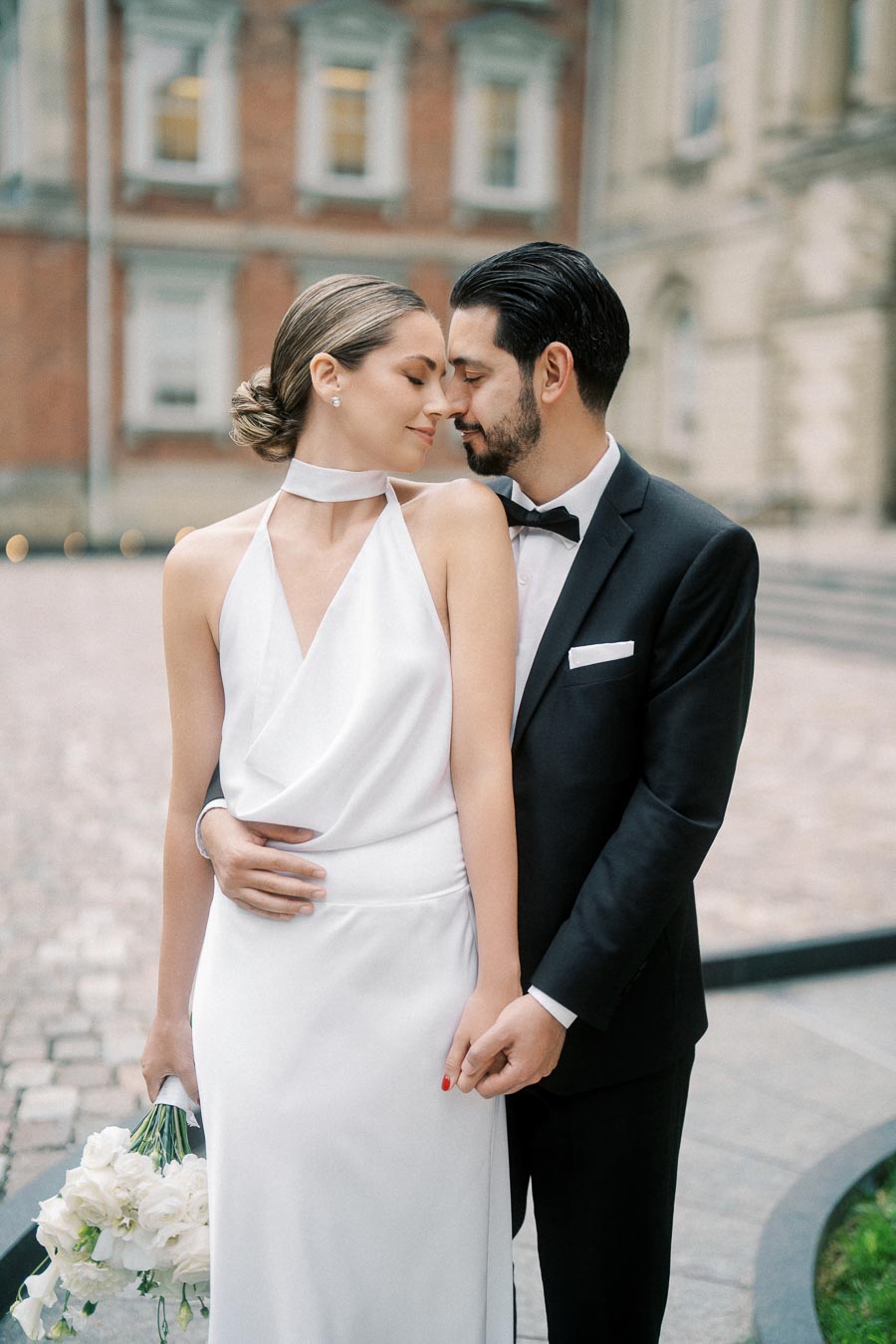 A bride in a white dress and groom in a black tuxedo embrace lovingly outdoors with a historic building in the background, highlighting their elegant wedding day.