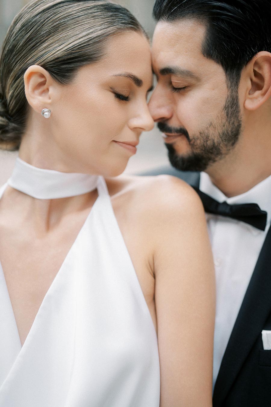 Elegant couple sharing an intimate moment, woman in white dress with pearl earrings and man in formal black suit and bow tie, showcasing romance and style.