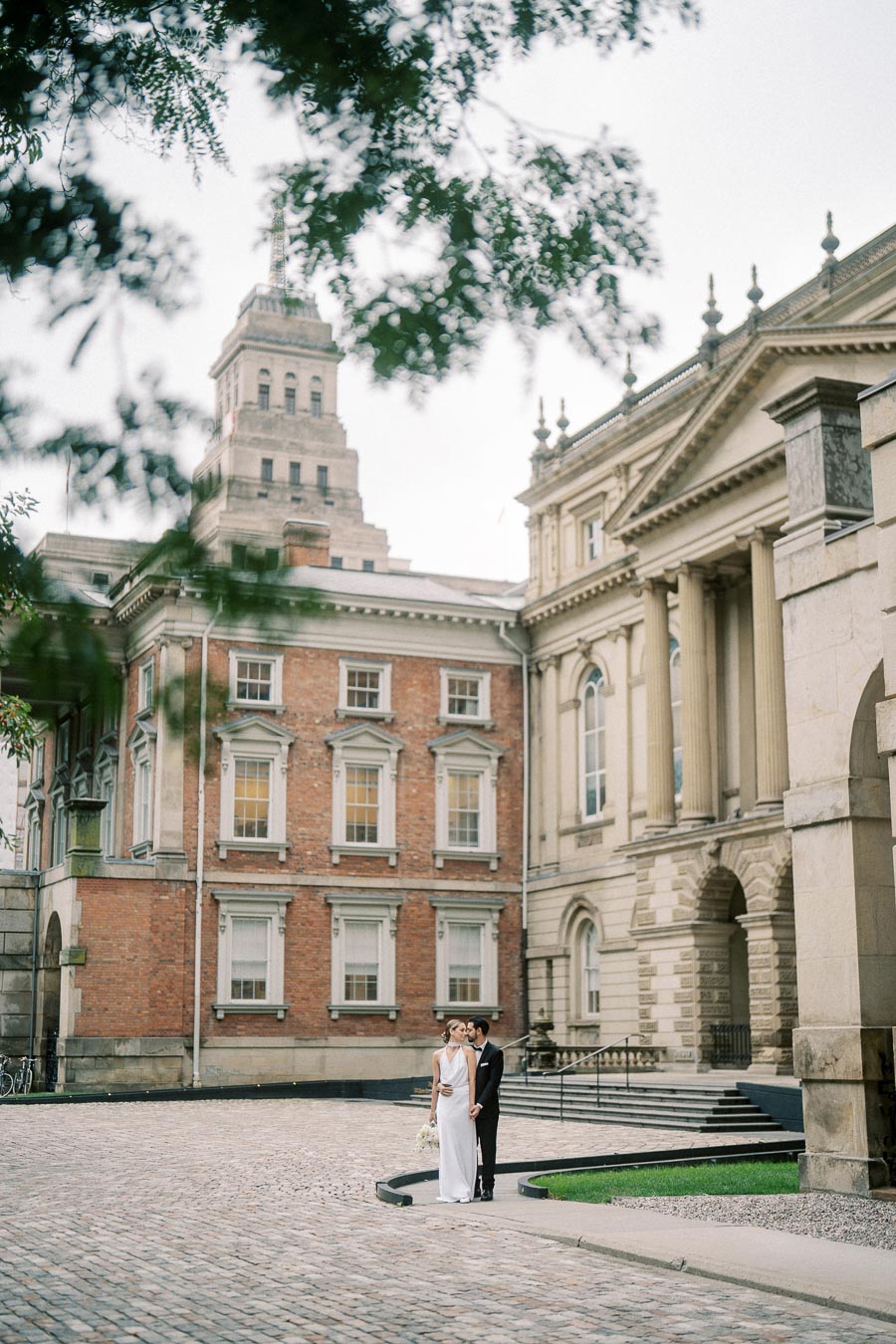Elegant couple in wedding attire standing on cobblestone pathway near historic brick and stone architecture with lush greenery, showcasing a picturesque urban wedding setting.