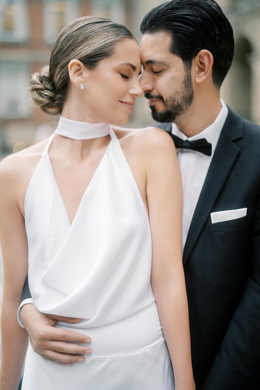 Romantic couple embracing in elegant attire, with the woman in a white gown and the man in a dark suit with a bow tie, sharing an intimate moment outdoors.