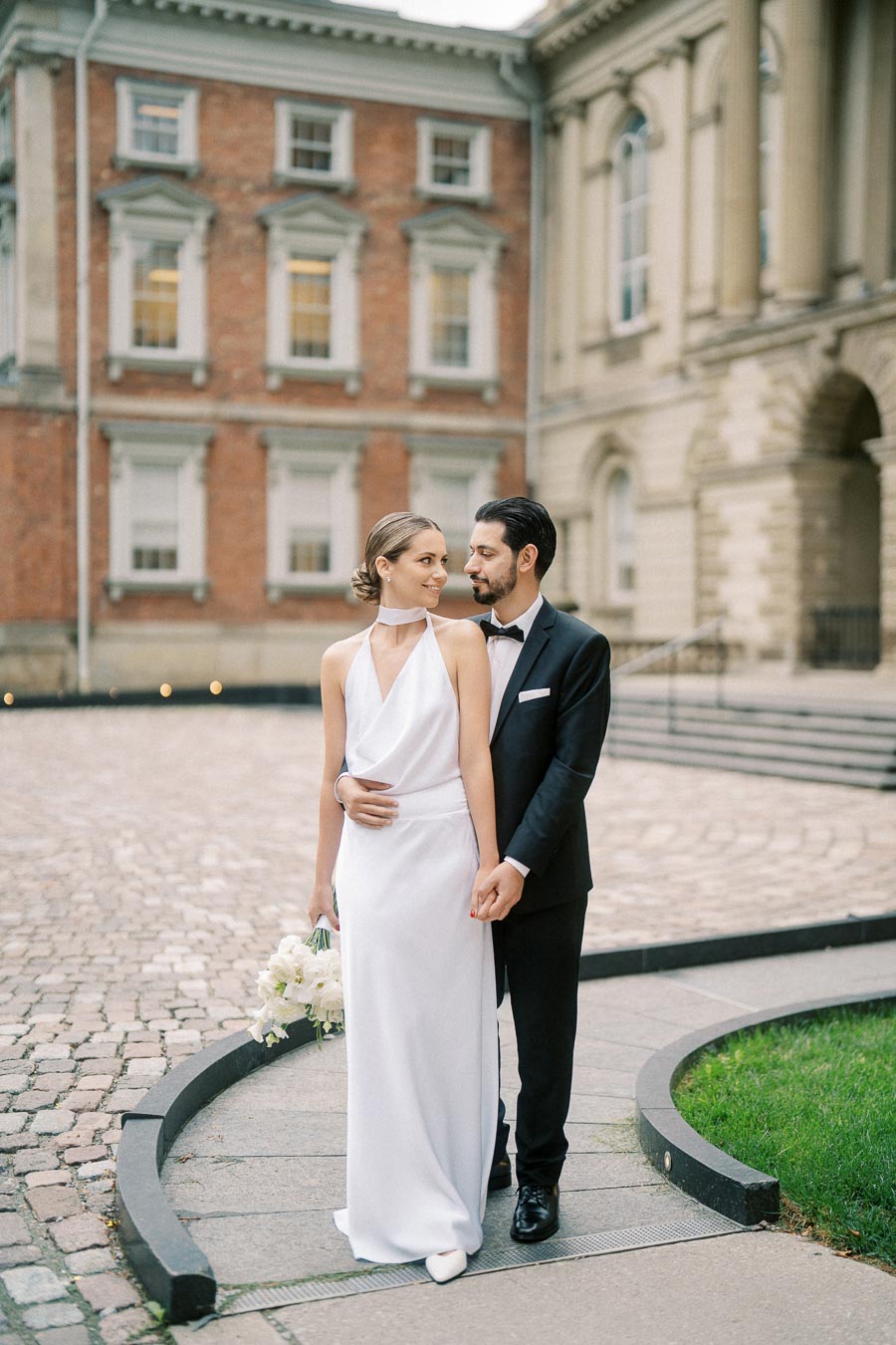 A bride in a white dress holding a bouquet stands with her groom in a black suit, posing in front of a historic building with red brick and classical architecture.
