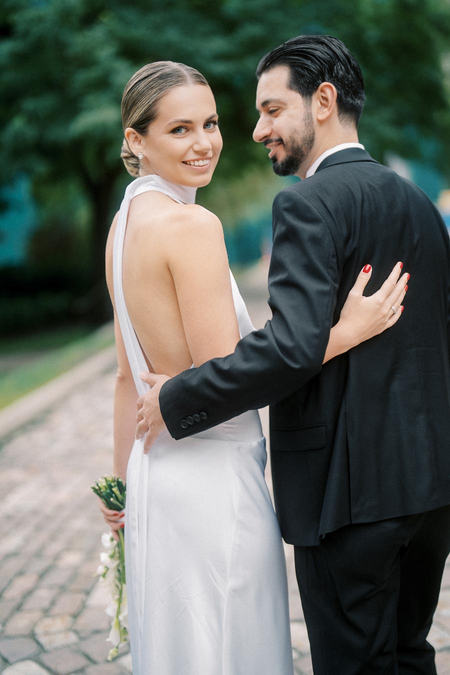Bride and groom embracing on a cobblestone path, with the bride in a sleek backless white dress and the groom in a black suit, capturing a joyful moment on their wedding day.