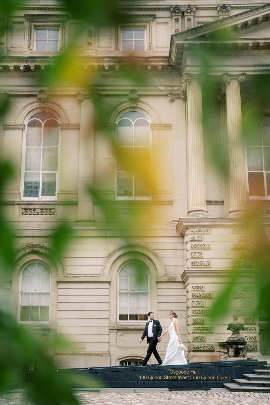 A newlywed couple walks hand in hand outside the historic Osgoode Hall, adorned with classical architecture and framed by lush greenery.