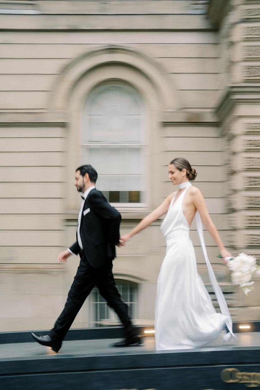 A bride in a flowing white dress holding a bouquet walks hand-in-hand with a groom in a black tuxedo, captured in motion against an ornate building backdrop.