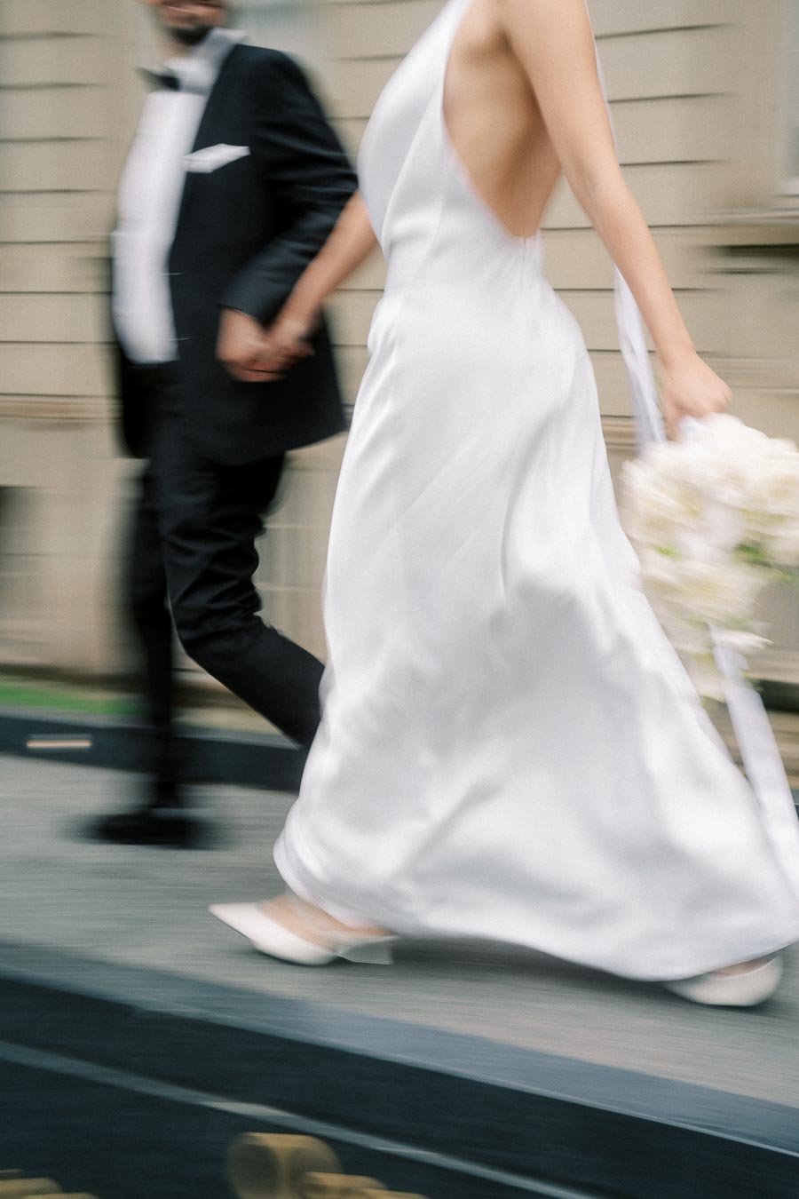 A bride in a flowing white dress walks hand in hand with a groom in a black tuxedo, each in motion, capturing the energy and emotion of a wedding day.