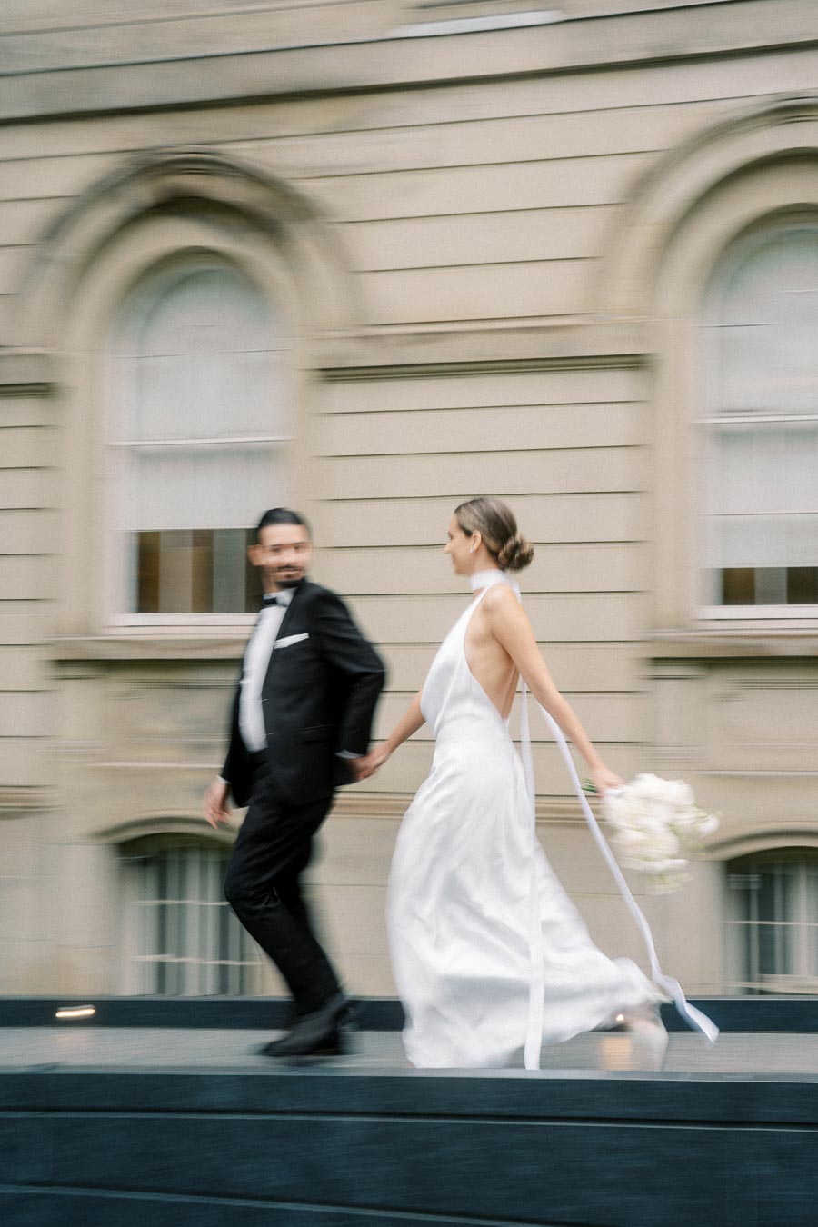 Blurry motion photograph of a newlywed couple holding hands, the bride in a flowing white gown carrying a bouquet, walking in front of an elegant historic building with arched windows.