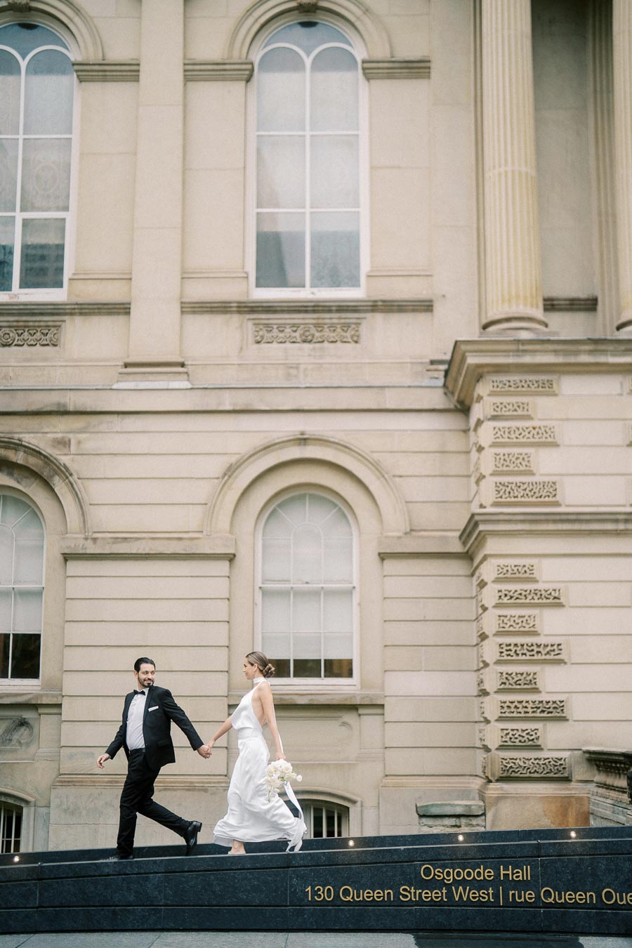 A couple in formal attire walking hand in hand in front of Osgoode Hall, Toronto. The bride holds a bouquet of white flowers, and the groom is in a black suit. The architectural backdrop features large windows and ornate detailing.