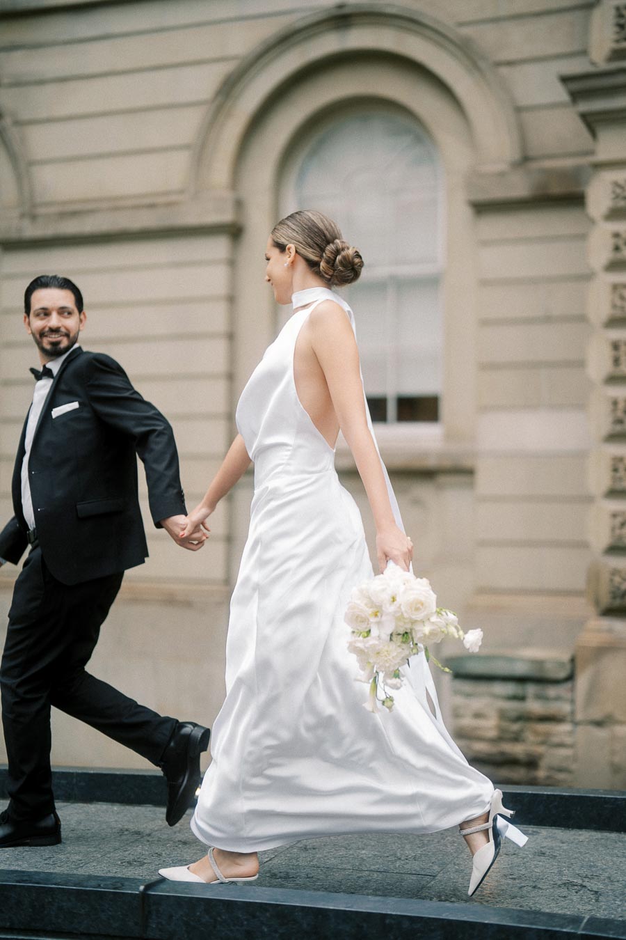 Elegant couple holding hands, bride in white gown carrying bouquet, walking outside historical building.