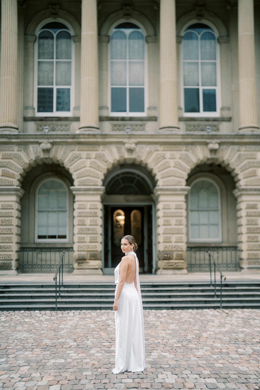 A woman in an elegant white gown stands in front of a historic building with arched windows and decorative columns.