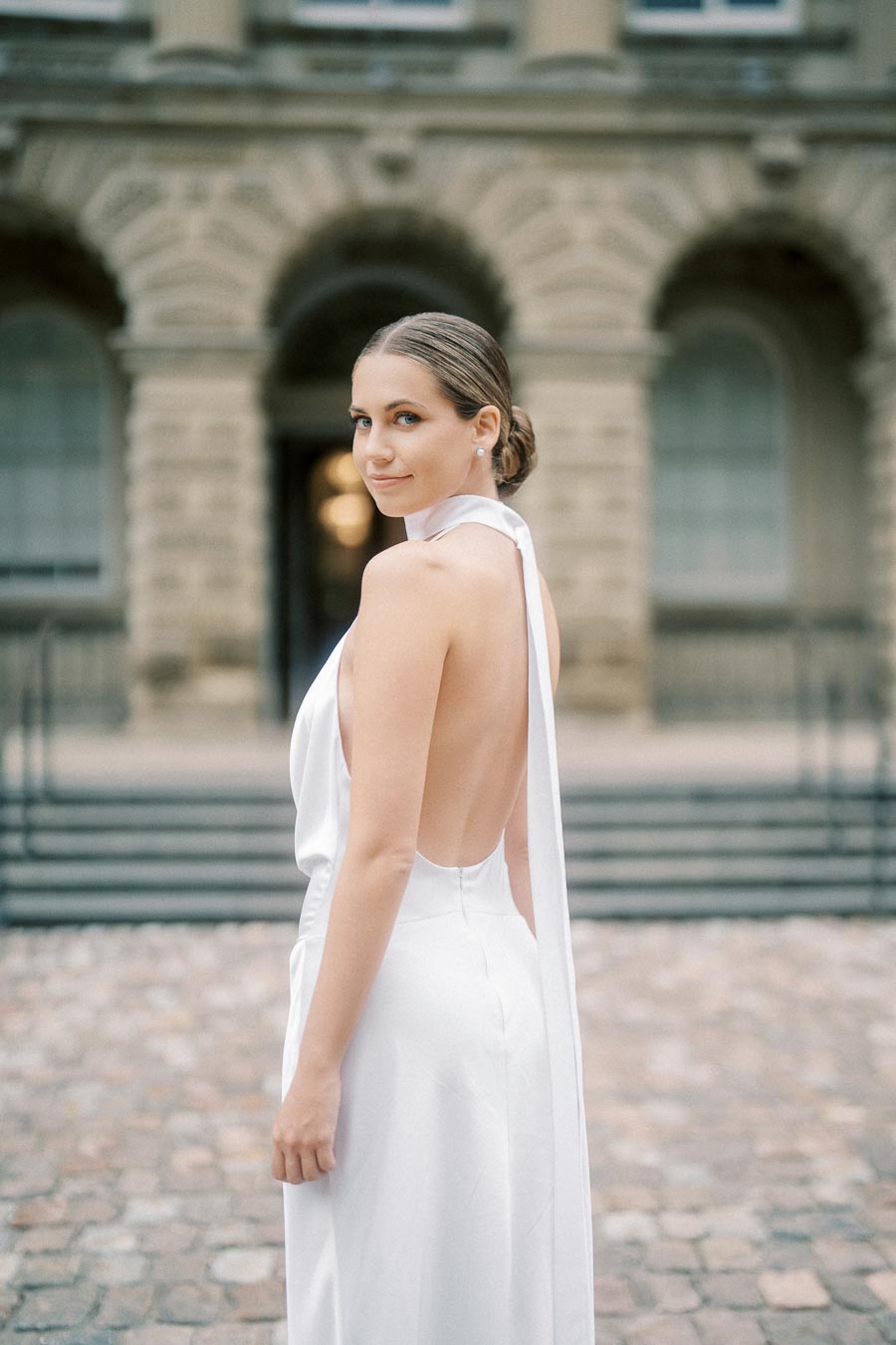 Elegant woman in a backless white dress standing outdoors in front of a historical building, showcasing classic wedding fashion and sophisticated style.