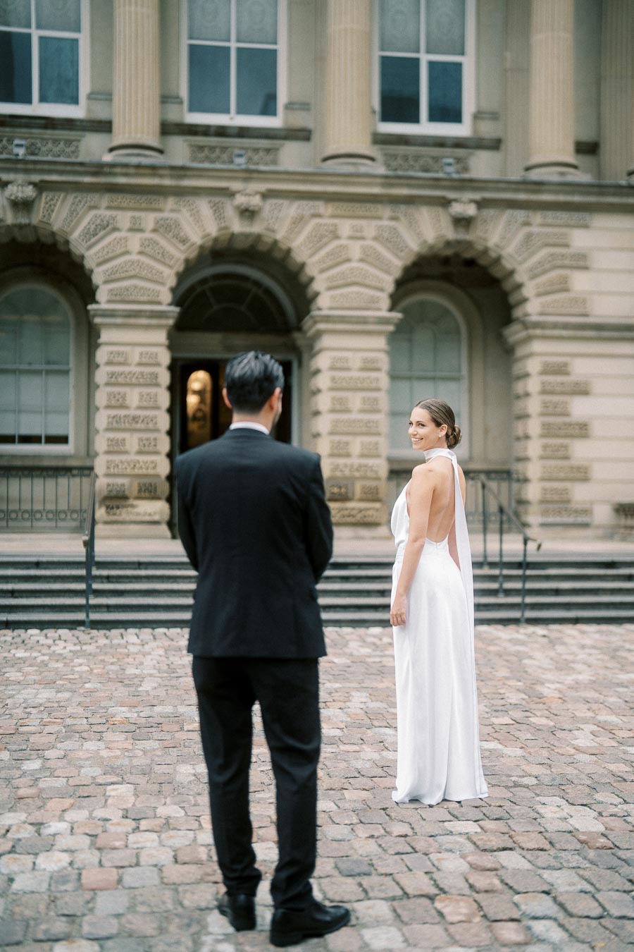 A bride in a white gown looks over her shoulder at a groom in a black suit, standing on a cobblestone path in front of a historic building with ornate columns.