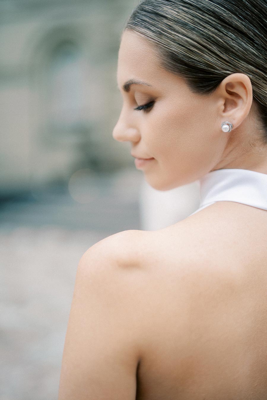 A woman with elegant hairstyle and pearl earrings, wearing a white halter dress, looks downwards in a serene outdoor setting.