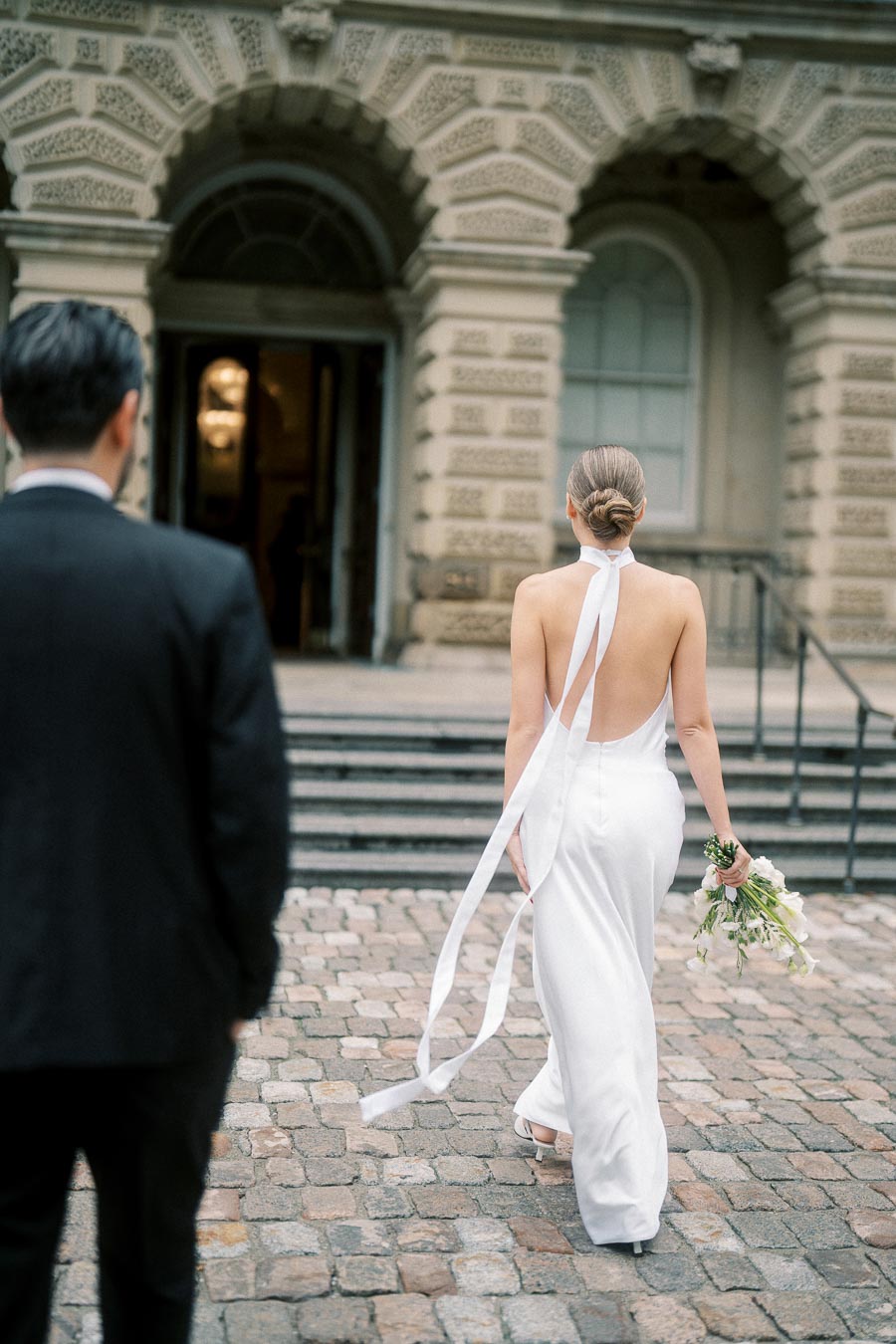 Bride in elegant white dress holding bouquet, walking towards historic building entrance, followed by a man in a suit on cobblestone path.