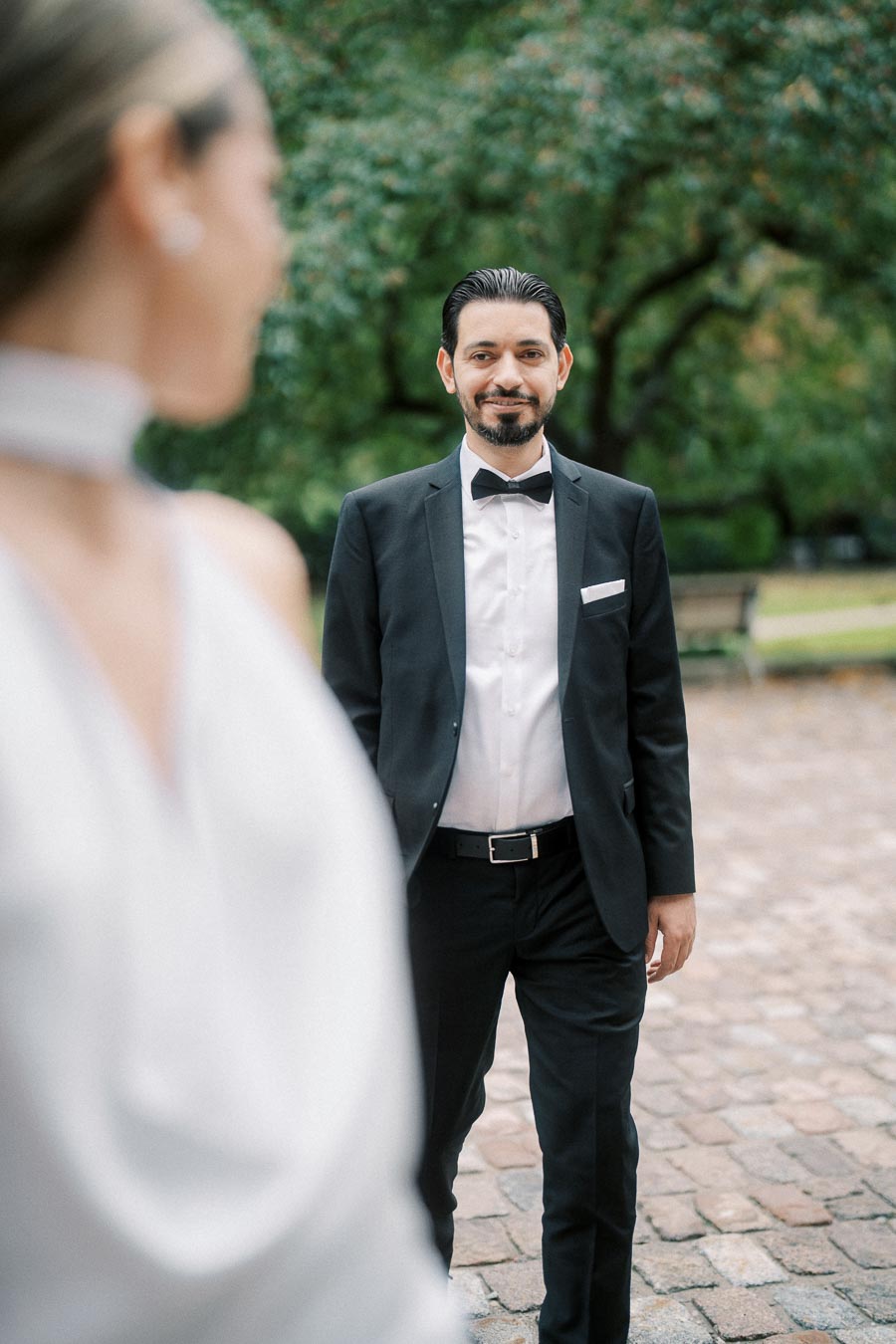 A man in a tuxedo smiles at a woman in a blurred white dress in an outdoor setting with lush greenery.
