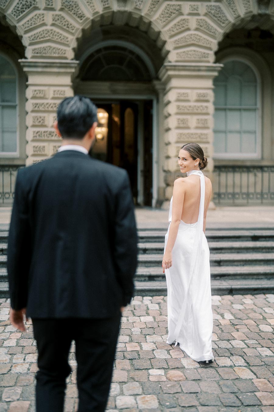 A bride in an elegant white gown smiles back at a man in a black suit, standing on cobblestone steps in front of a historic building.