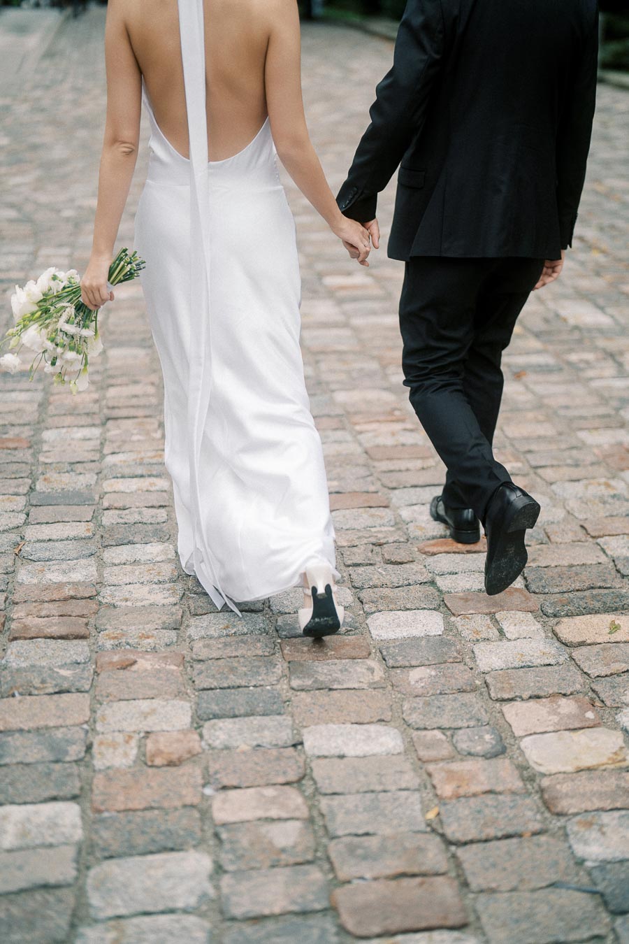 A couple holding hands, walking on a cobblestone path. The woman is in a backless white wedding dress, carrying a bouquet, and the man is in a black suit.