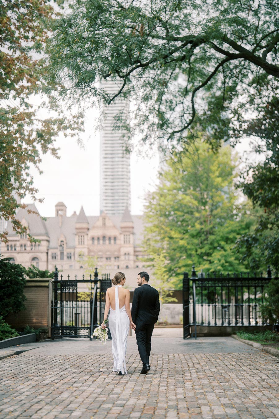 A bride in a white dress and a groom in a black suit walk hand in hand on a cobblestone path, surrounded by lush trees and historic architecture in the background.