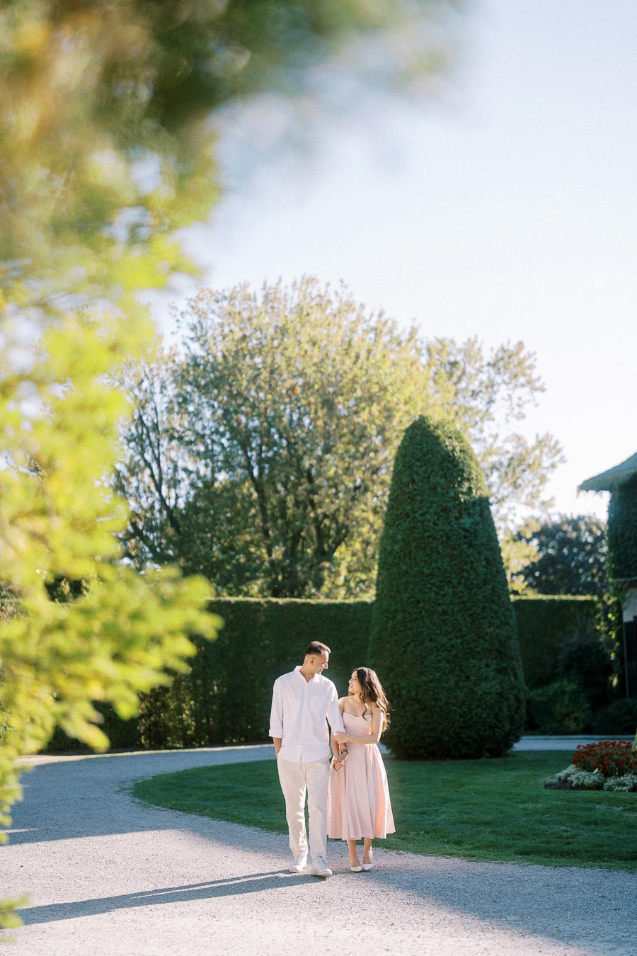 A couple walks hand in hand along a sunlit garden path lined with manicured hedges and lush greenery, creating a romantic and serene atmosphere.
