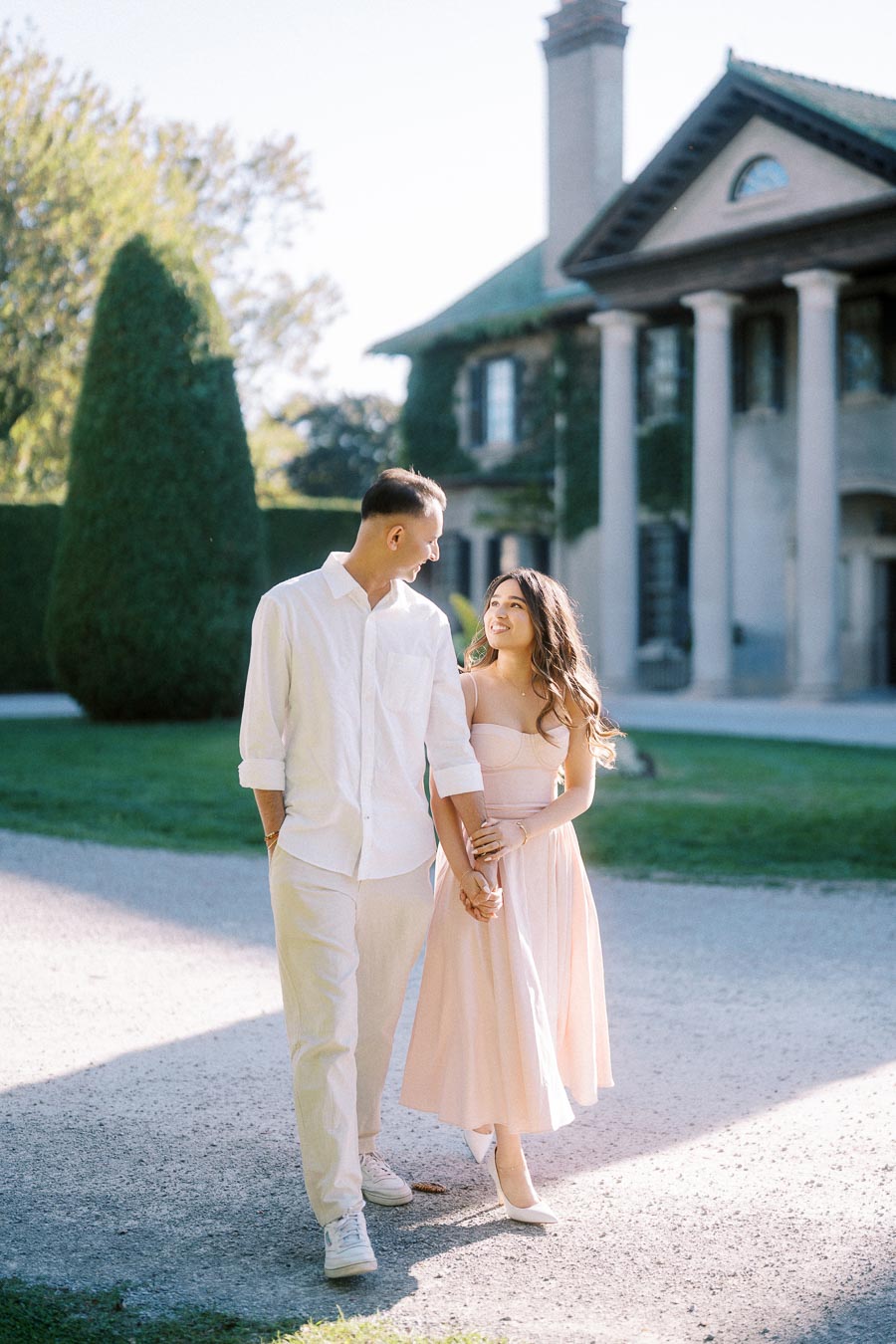 A couple walking hand in hand in front of a historic mansion on a sunny day, surrounded by lush greenery.