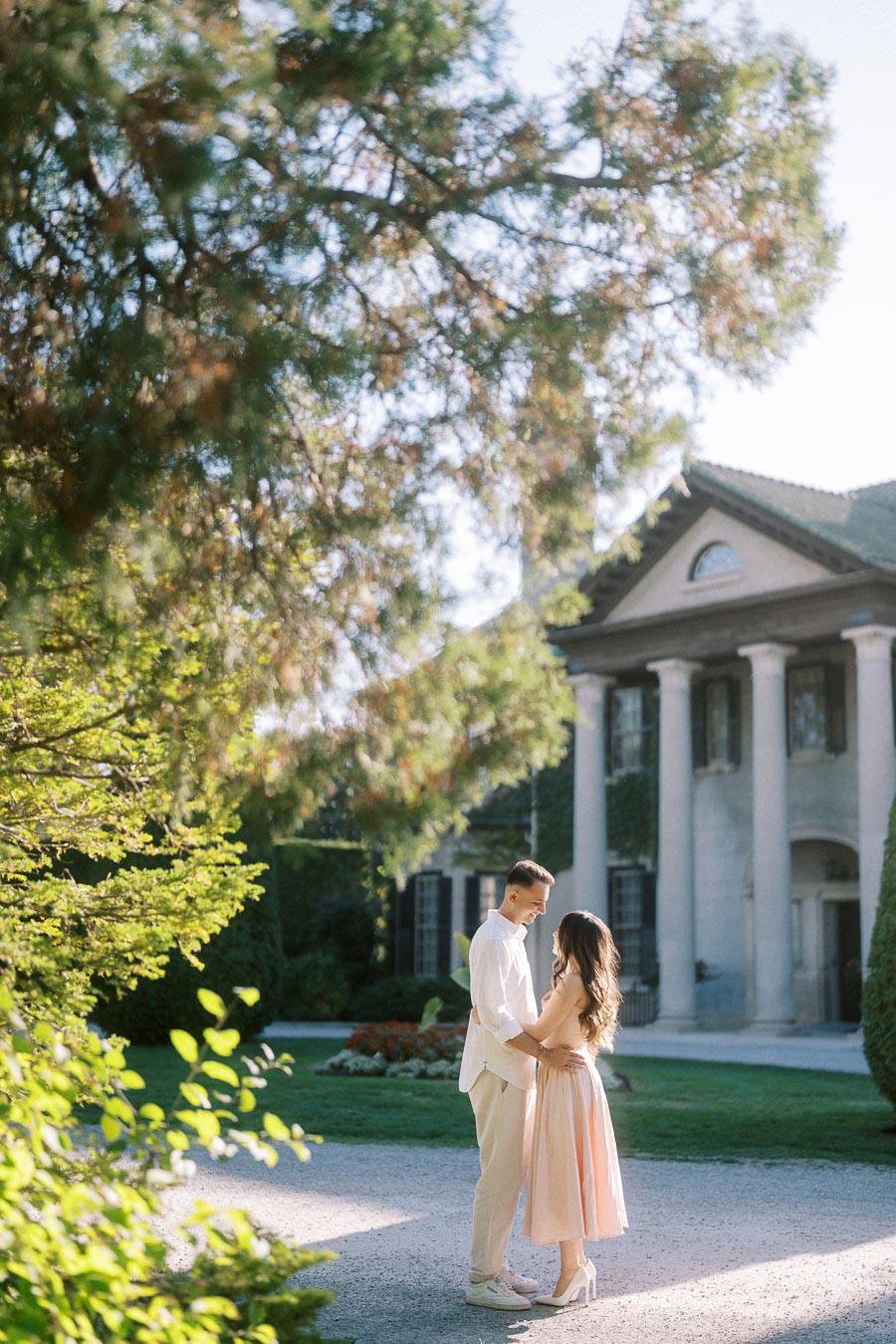 A couple embracing in front of a grand, historic mansion surrounded by lush greenery and bright sunlight, creating a romantic and elegant scene.