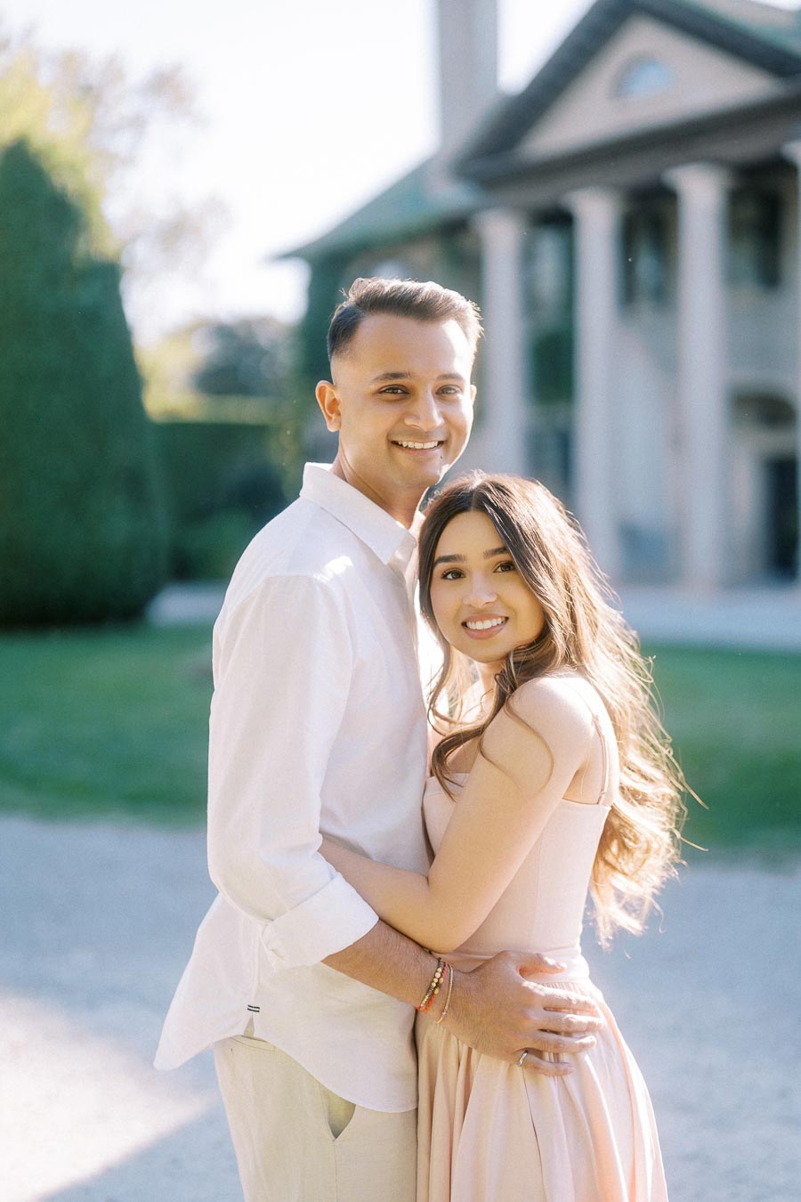 Happy couple embracing outdoors in front of an elegant building, dressed in light pastel attire under natural sunlight.