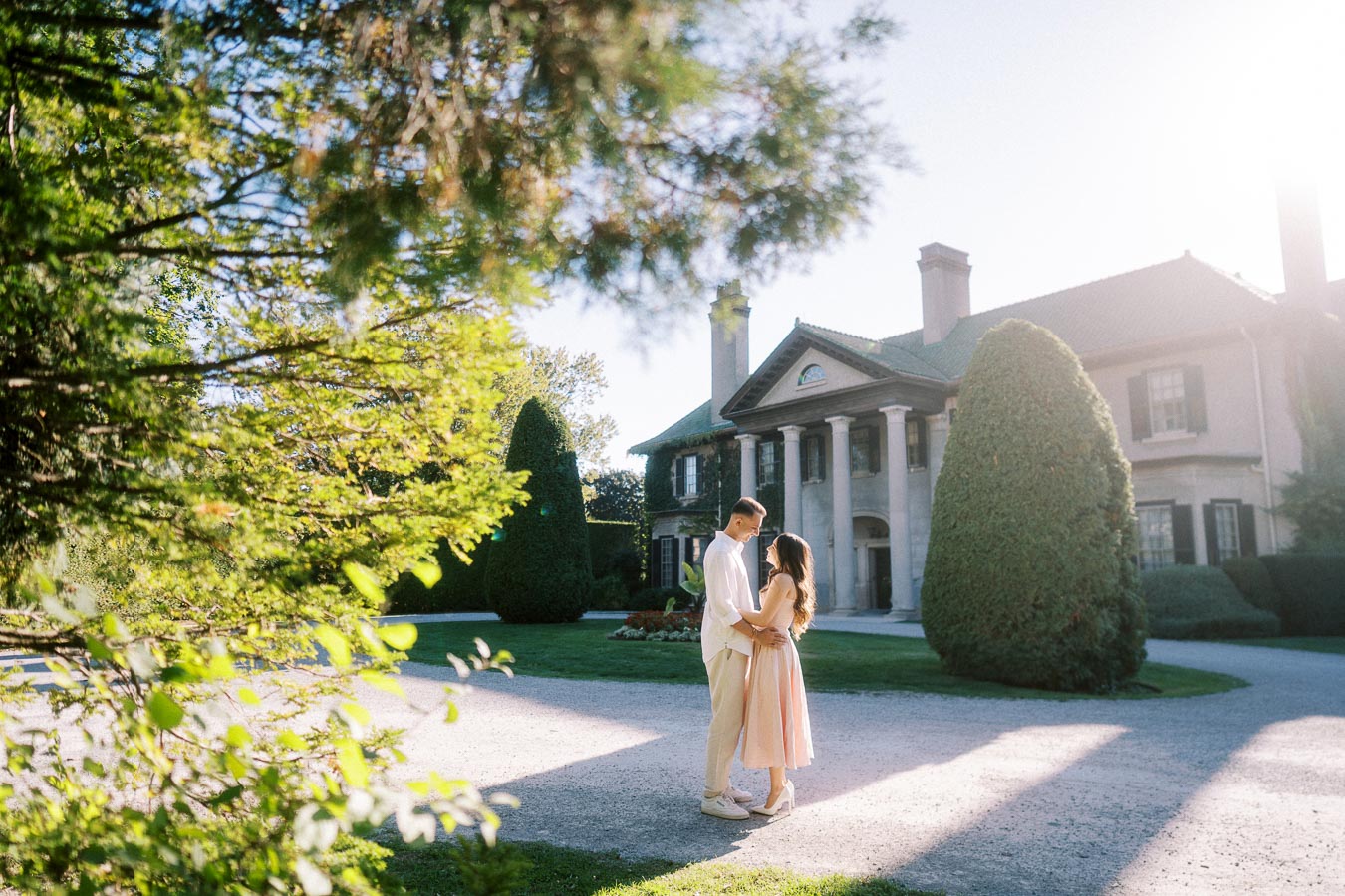 A couple embracing in front of an elegant, historic mansion surrounded by lush greenery and bright sunlight.
