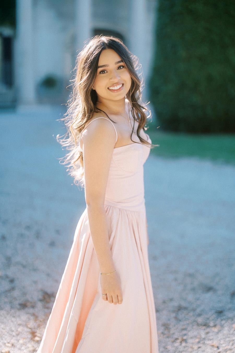 Young woman in a soft peach gown smiling outdoors, with sunlight enhancing her hair, standing on a gravel path with greenery in the background.