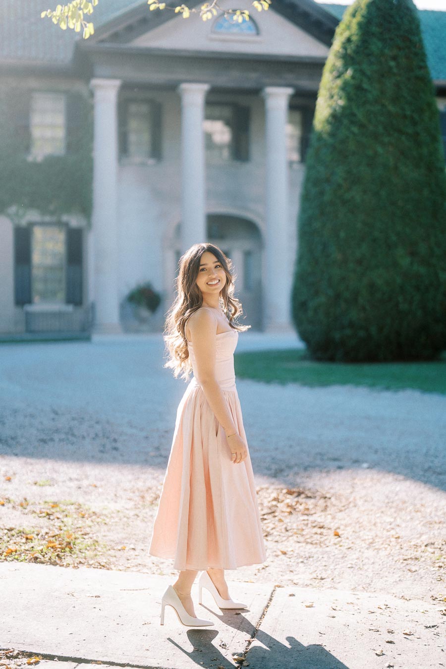 Young woman in a blush pink dress and high heels smiling in front of a grand mansion with large columns and a well-maintained garden under soft sunlight.
