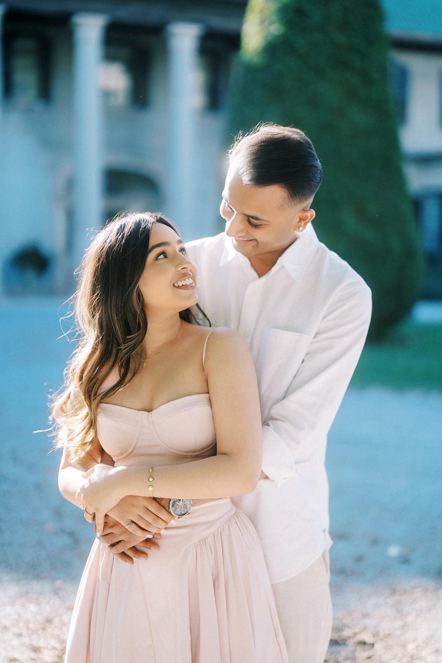 A couple embraces lovingly in front of a picturesque building, with the woman wearing a light pink dress and the man in a white shirt, both smiling at each other against a soft background of green foliage and classic architecture.