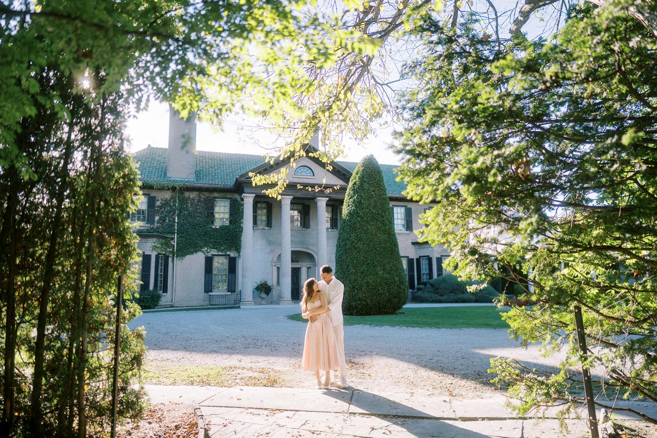 Elegant couple embracing in sunlight in front of a historic mansion surrounded by lush greenery, capturing a romantic outdoor moment.