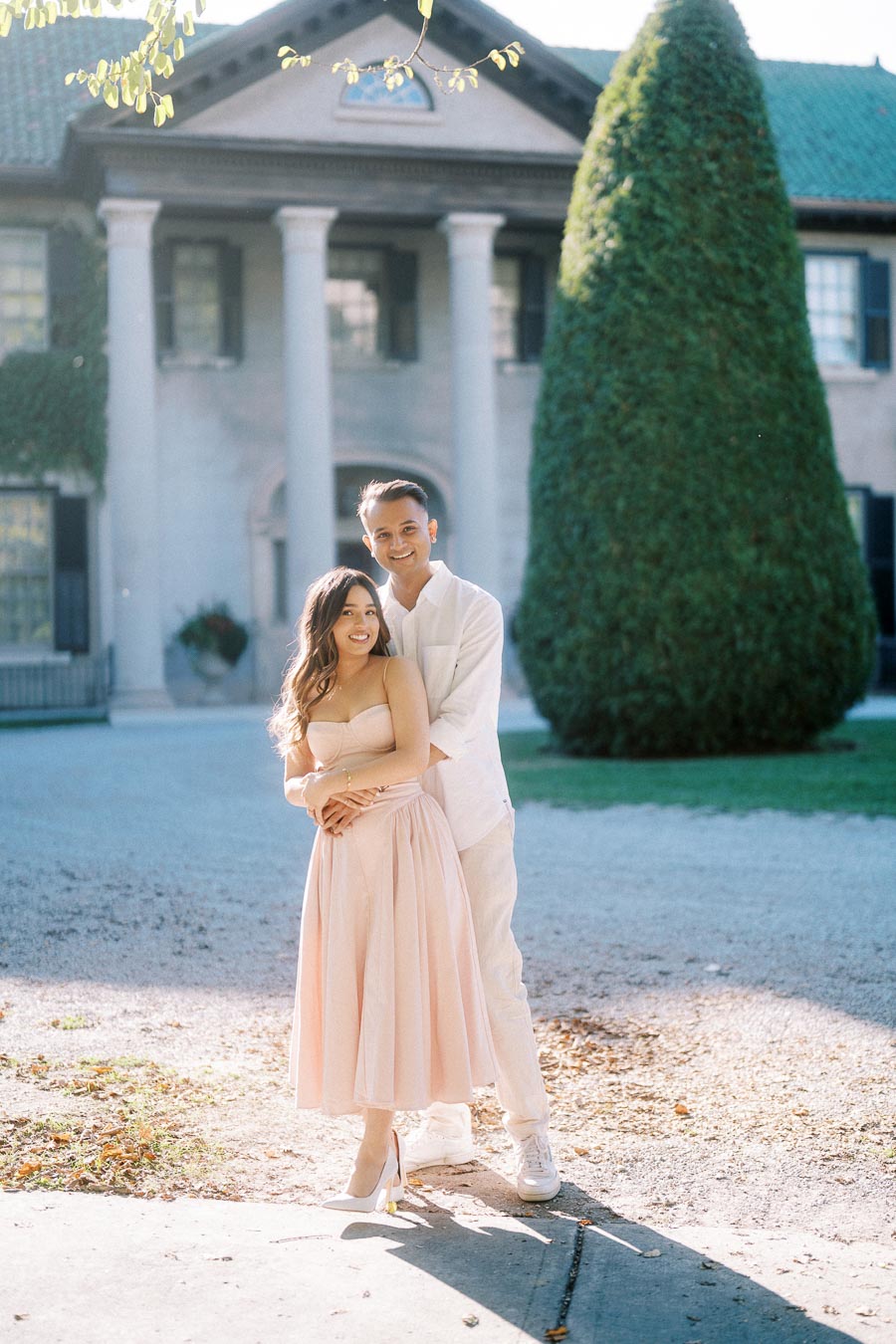 A couple embracing in front of a historic mansion, with the woman in a pastel pink dress and the man in a white outfit, surrounded by natural greenery.