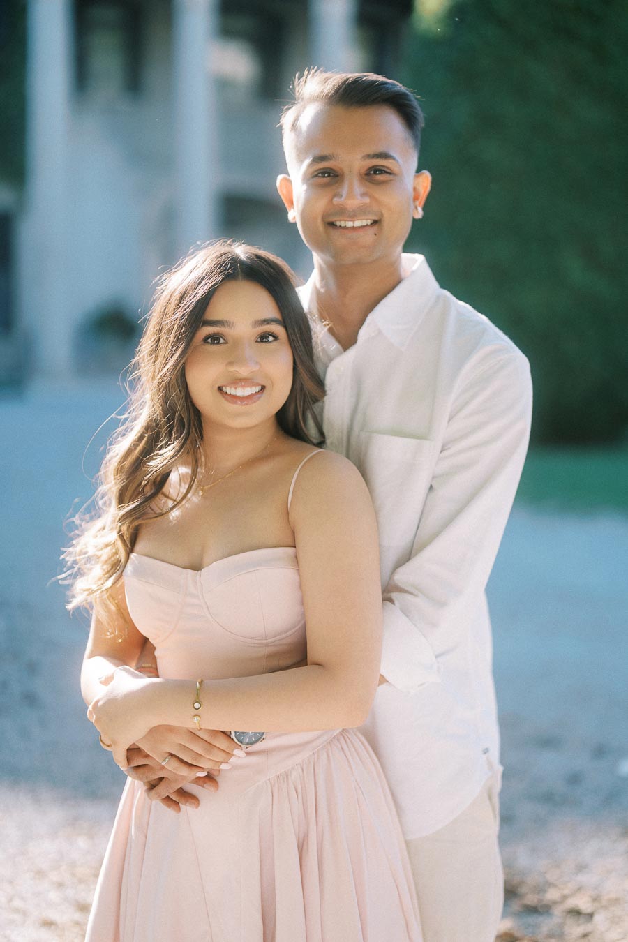 A smiling couple embracing each other outdoors, with soft sunlight highlighting their expressions. The woman is wearing a light pink dress and the man is in a white shirt, standing in a peaceful, natural setting.