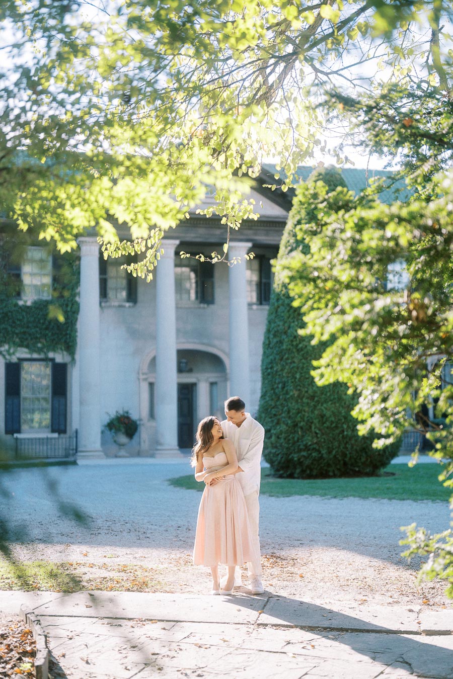 Romantic couple embracing in front of a grand historic mansion on a sunny day, surrounded by lush greenery and elegant architecture.