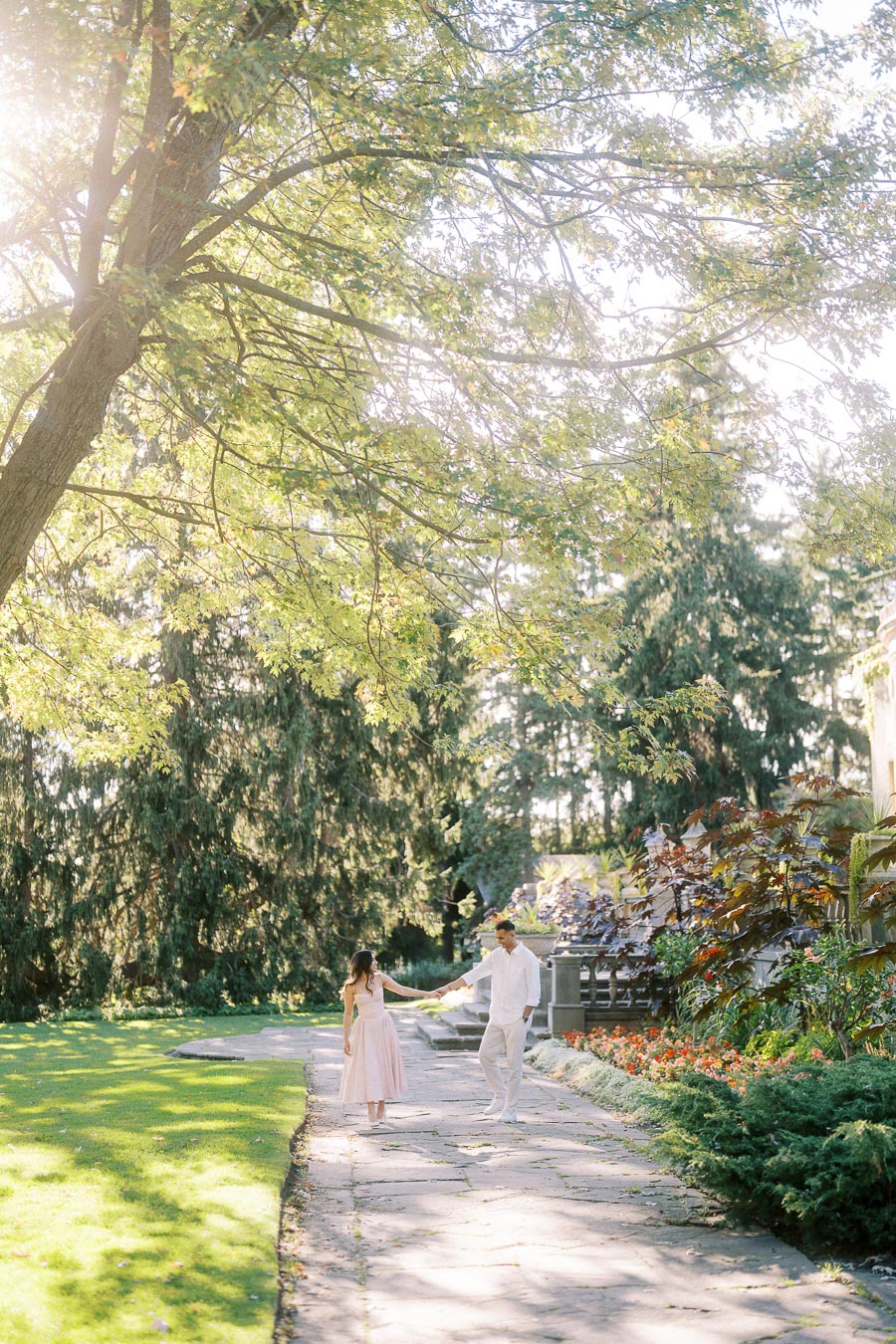 Young couple walking hand in hand along a sunlit garden path surrounded by lush greenery and colorful flowers.