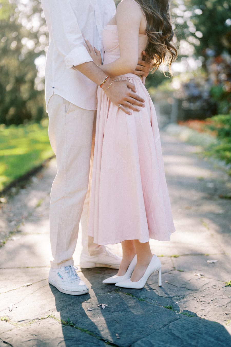 A couple standing close together on a sunlit garden path, with the focus on their lower bodies; the woman is wearing a pink dress and white heels, while the man is dressed in white pants and sneakers, surrounded by greenery.