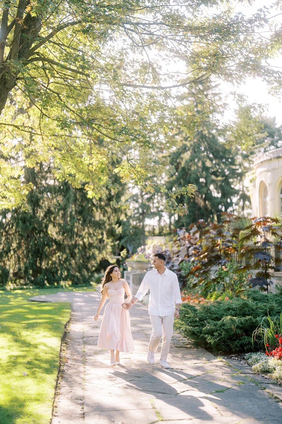 A happy couple strolling hand in hand along a sunlit garden path, surrounded by lush greenery and vibrant flowers.