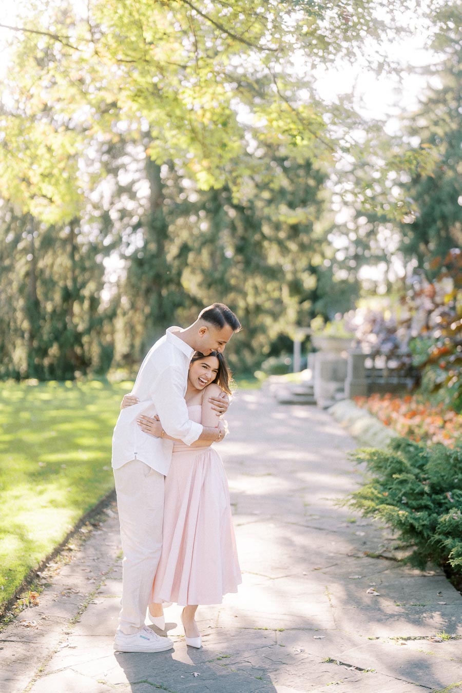 A couple embracing and smiling in a sunlit park, surrounded by lush greenery and flowers, enjoying a romantic moment on a stone pathway.