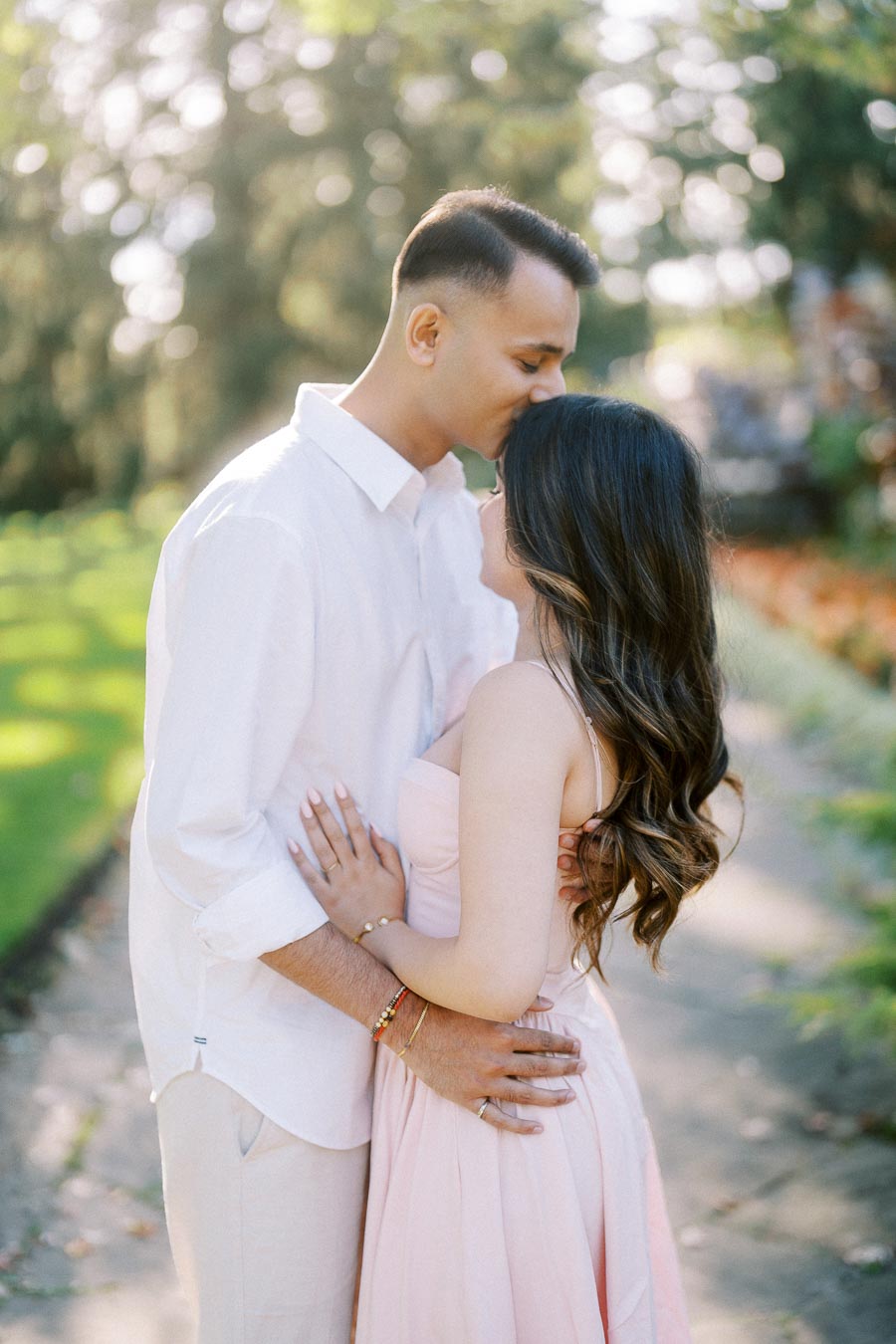 Couple sharing an intimate moment in sunlight, man gently kissing woman's forehead in a serene garden setting.